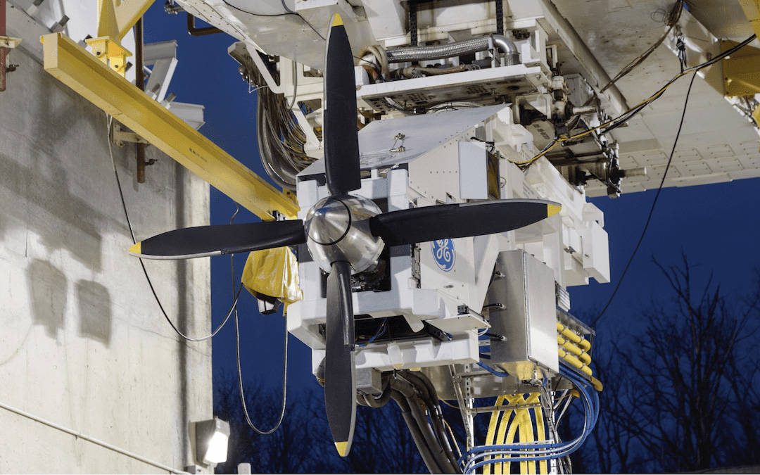 A close-up view of a propeller system mounted on a testing rig. The propeller has four black blades and is attached to a white mechanical assembly. The background features industrial equipment and a dark evening sky.