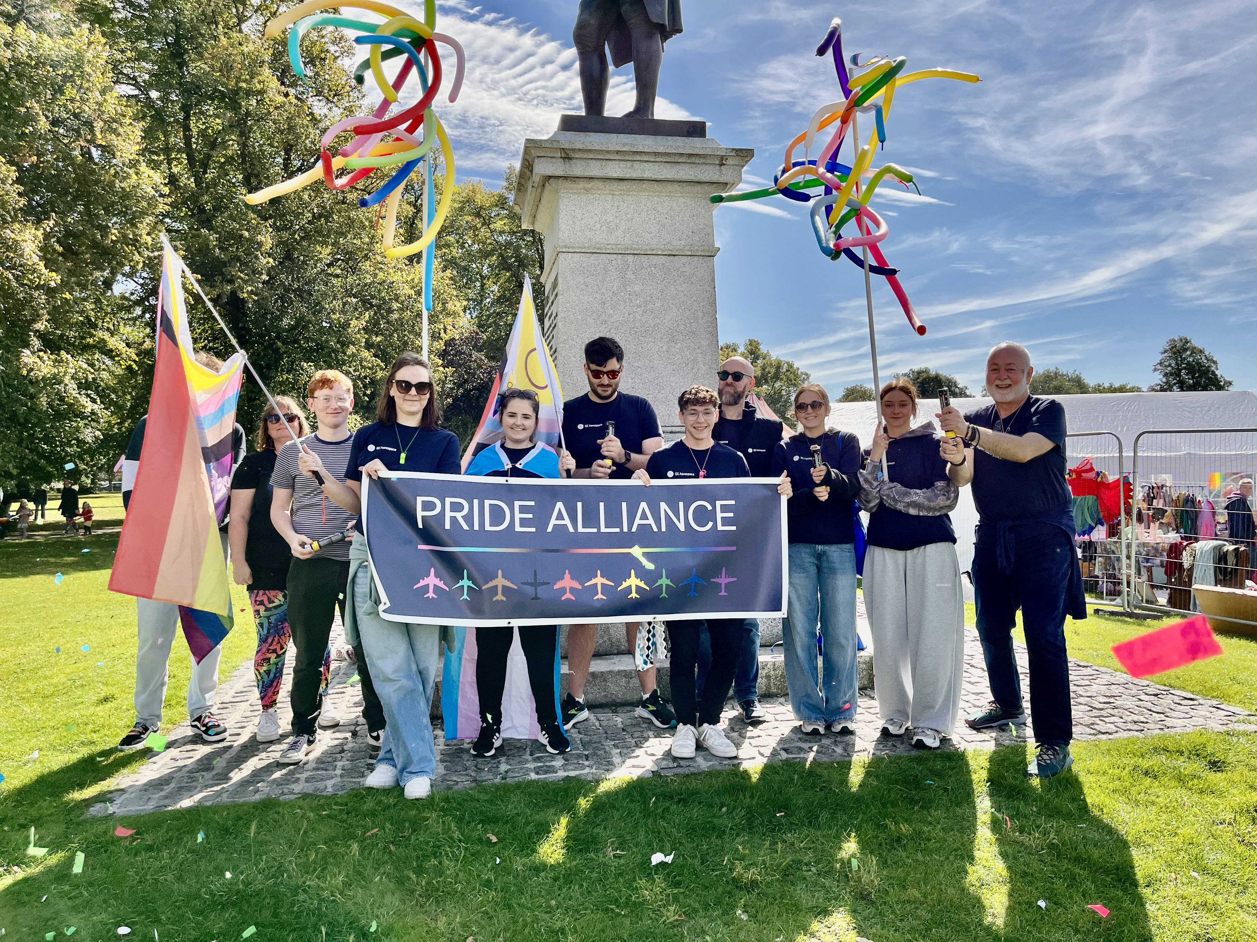 The GE Aerospace Pride Alliance members gather for a local Pride parade, holding a Pride Alliance banner in front of a statue and waving balloon creations.