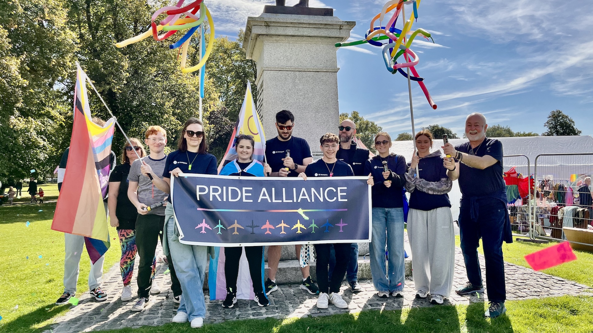 The GE Aerospace Pride Alliance members gather for a local Pride parade, holding a Pride Alliance banner in front of a statue and waving balloon creations.