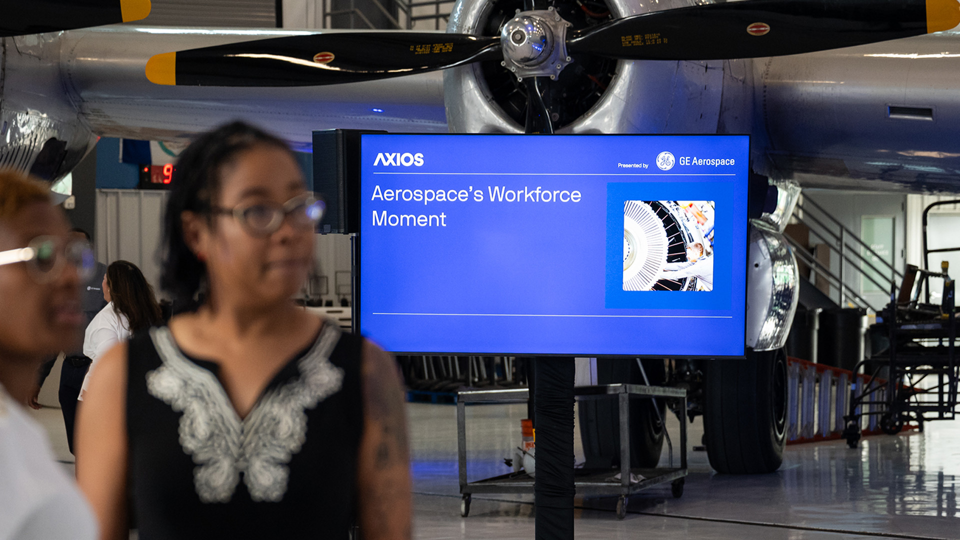Event screen in an aircraft hangar reading ‘Aerospace’s Workforce Moment,’ with Axios branding and ‘Presented by GE Aerospace.’ Attendees stand in the foreground beneath a vintage propeller plane.