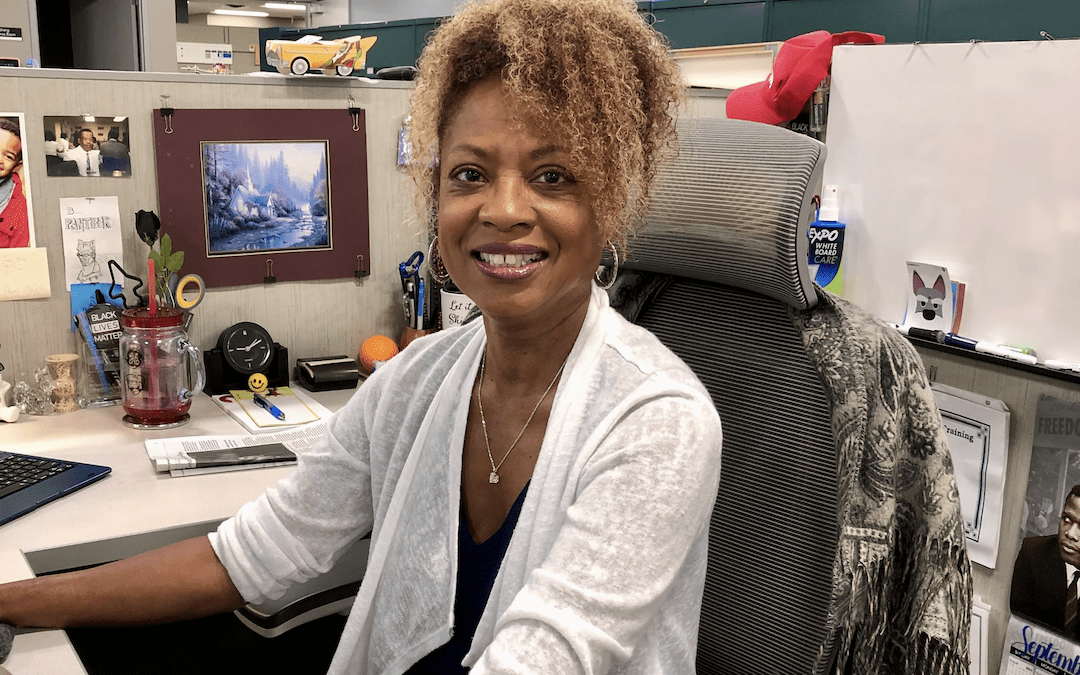 A person sitting at a desk in an office, wearing a white cardigan and a blue top.