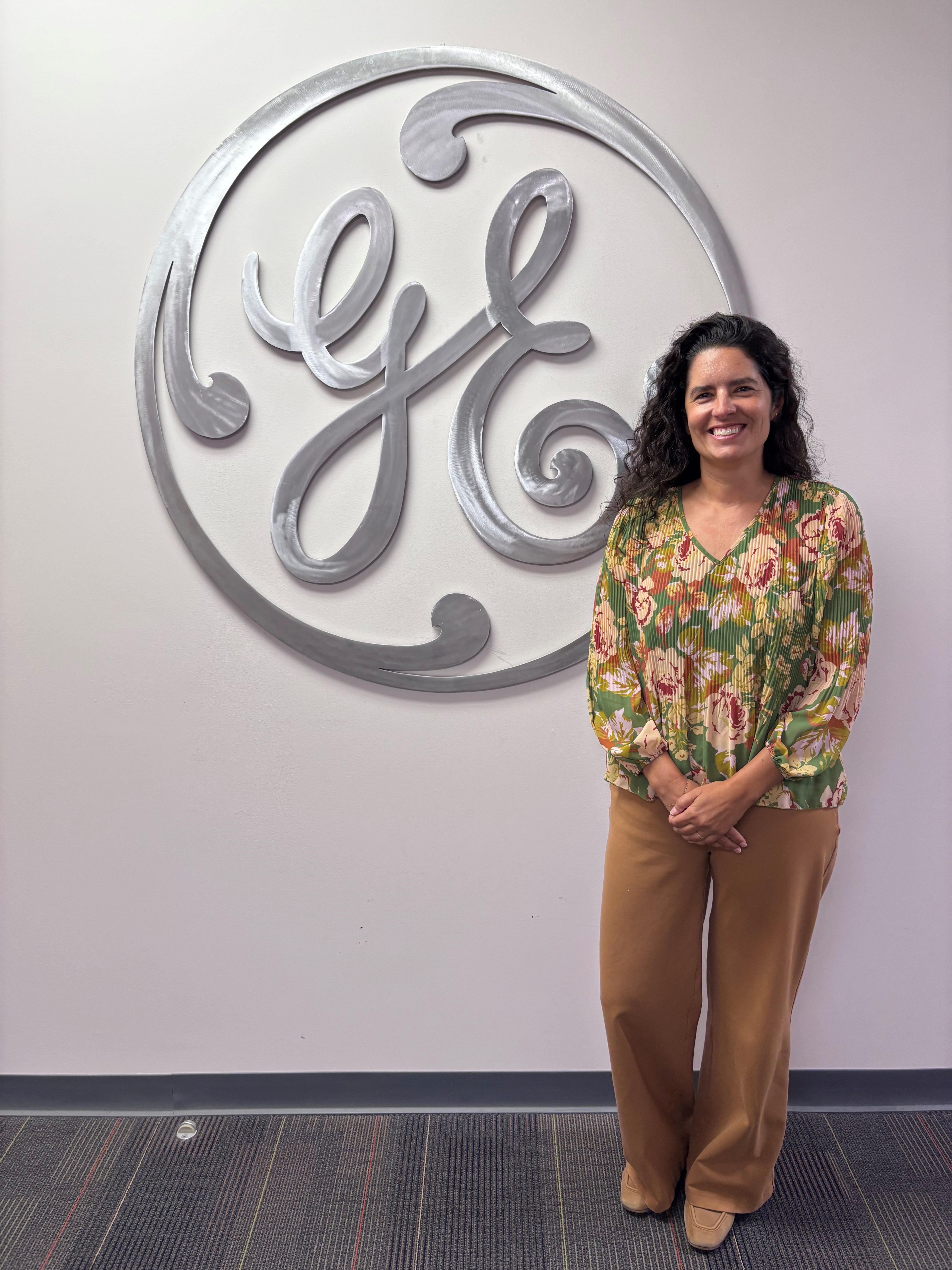 Gabrielle Boesenberg stands in a flowered top in front of a silver GE logo on a white wall