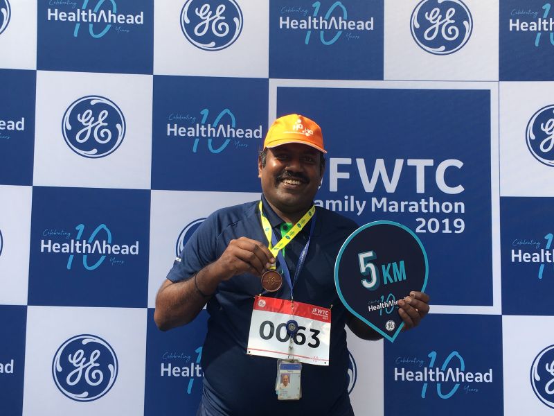 Man standing in front of a marathon backdrop holding a 5 km sign and holding up a medal
