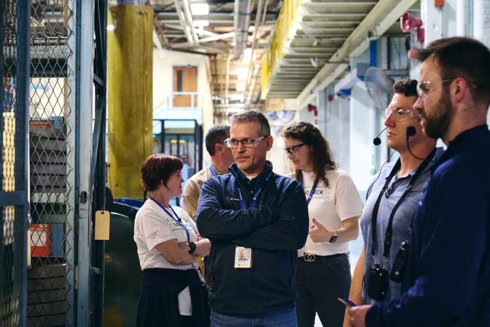 Adam Baran stands with his arms crossed near the heat-treat oven, with colleagues surrounding him looking on