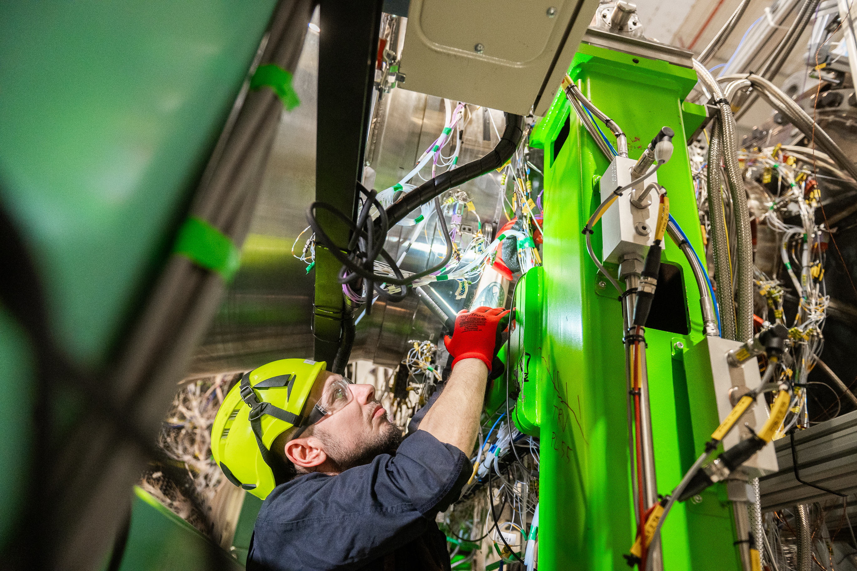 Technician in a hard hat and safety glasses reaches up to work on a bright green...