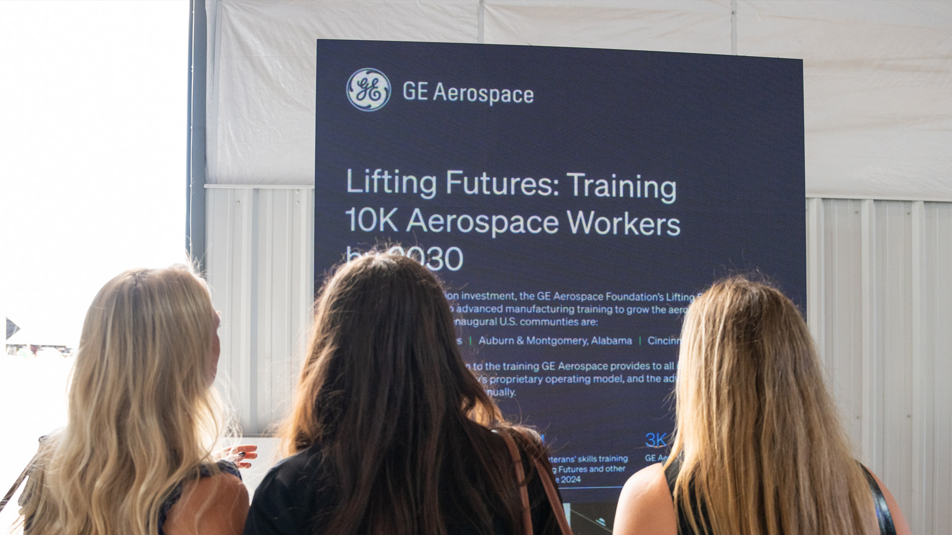 Three women with long hair, photographed from behind, look at a large display reading “Lifting Futures: Training 10K Aerospace Workers by 2030” under the GE Aerospace logo.